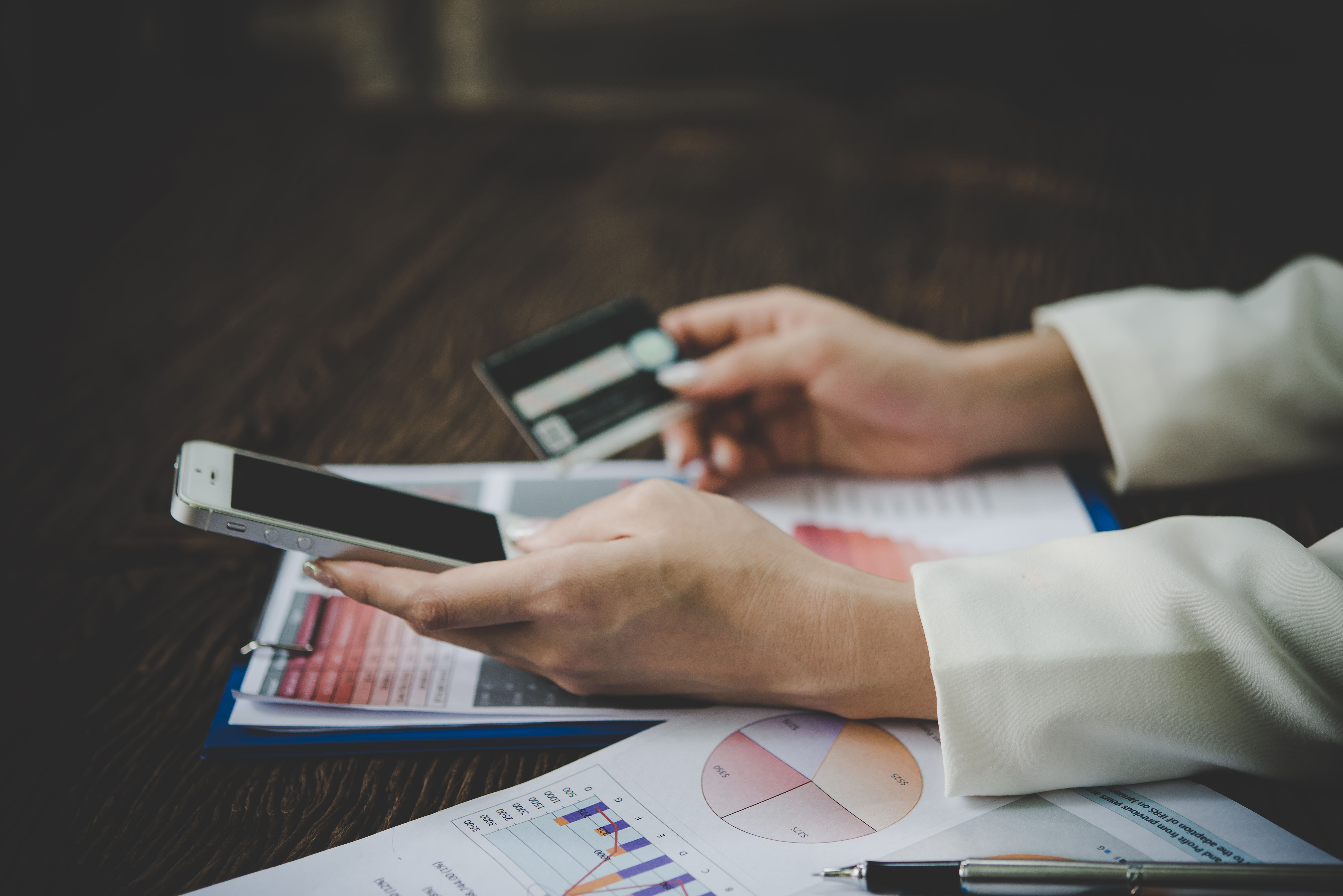 Hands holding smartphone and credit card over printed financial charts — custom software 				for accounts receivable and payable management.